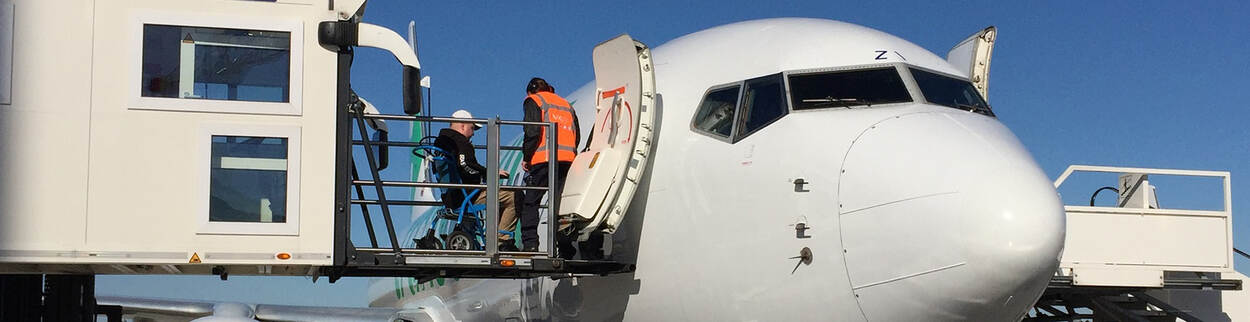 Male passenger in wheelchair is assisted by airport staff in boarding aeroplane