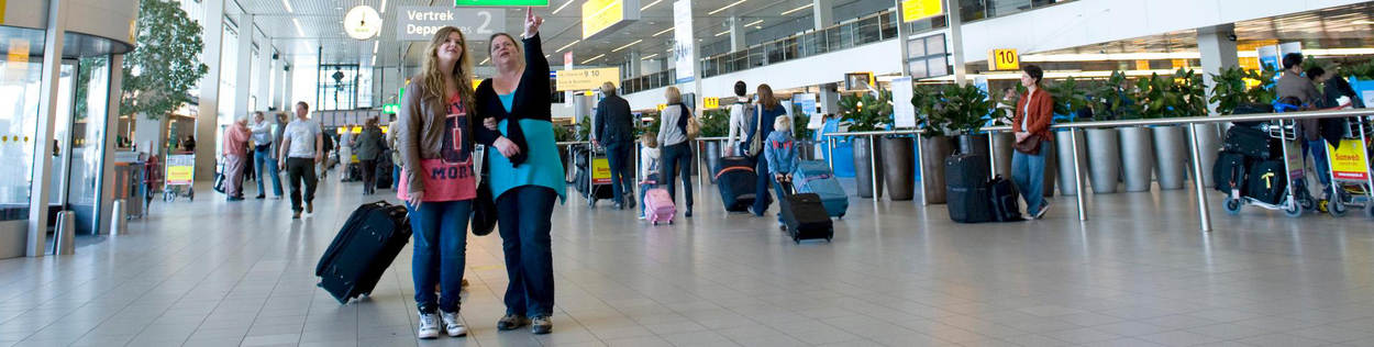 In de vertrekhal van luchthaven Schiphol kijken twee vrouwen naar een bord met informatie. Een van hen heeft een koffer. Op de achtergrond wandelen mensen met baggage.